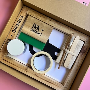May include: A screen printing kit in a cardboard box. The kit includes a wooden frame, a green squeegee, a roll of tape, a white container, and a small bag with tools. The box has the text "INK + ORANGES".