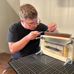 May include: A wooden bookbinding press featuring four threaded metal rods with wing nuts at each corner, a central handle, and a rectangular wooden frame. The press is used to hold a stack of paper sheets securely in place during the binding process.