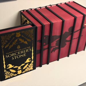 May include: A set of seven hardcover books with black covers and gold foil detailing. The front book is titled Harry Potter and the Sorcerer's Stone. The remaining six books feature red spines that form a continuous silhouette of a figure on a broom.