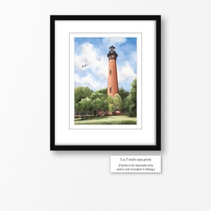 May include: Watercolor painting of a red brick lighthouse with a white trim and a black roof. The lighthouse is surrounded by green trees and a blue sky with white clouds. The text "5 x 7-inch size print (Frame is for example only and is not included in listing.)" is at the bottom of the image.