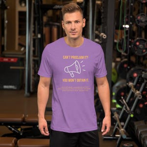 Puede incluir: Camiseta de manga corta de color morado con un contorno blanco de un megáfono y el texto "Can't proclaim it? You won't obtain it." en amarillo. El texto "With the heart a man believeth unto righteousness; and with the mouth confession is made unto salvation." está impreso en amarillo más pequeño debajo del megáfono.