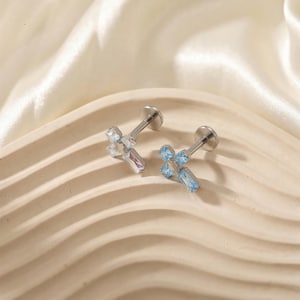 May include: Two silver-toned cross-shaped earrings with iridescent and light blue gemstones. The earrings are displayed on a beige, wavy surface, with a white, silky fabric in the background. The gemstones are cut in various shapes.