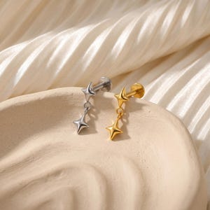 May include: Two star-shaped earrings, one silver and one gold, displayed on a beige surface. Each earring features a small star stud and a dangling star charm. The earrings are designed for ear piercings.