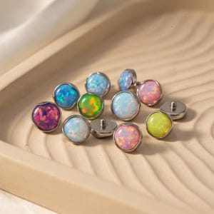 May include: A collection of opal cabochons in various colors, including blue, pink, green, and white, set in silver-colored metal. The opals are round and have a polished, iridescent appearance. They are displayed on a light-colored tray.