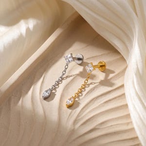 May include: Two stud earrings, one silver and one gold, each featuring a square-cut clear gemstone and a dangling teardrop-shaped gemstone. The earrings are connected by a delicate chain and are displayed on a neutral-toned surface.