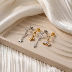 May include: Four pieces of jewellery, including three gold and silver-toned earrings. One earring features a dangling star and moon charm, another has a sunburst charm, and the third has a chain with a teardrop and a crown charm. The earrings are displayed on a light brown tray.