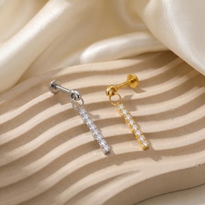 May include: Two ear piercing studs, one silver and one gold, each with a dangling bar of clear, sparkling stones. The studs are displayed on a beige, textured surface with a cream-colored fabric in the background.