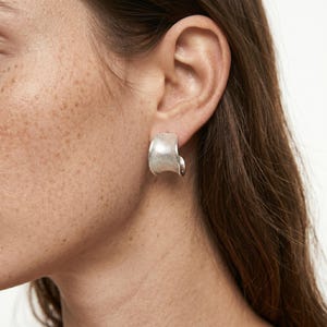 May include: A silver-toned, textured earring is shown on a person's ear. The earring has a curved, open design, with a slightly rough surface. The earring is a simple, yet elegant piece of jewelry.