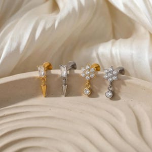 May include: Four stud earrings in gold and silver tones. Two have a rectangular gem and a dangling spike. The other two feature a flower design with clear gems and a dangling gem. The earrings are displayed on a beige surface.