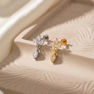 May include: Two delicate earrings, one silver-toned and one gold-toned, each featuring a cluster of small, clear crystals in a crown-like design. A teardrop-shaped crystal dangles from each earring. The earrings are displayed on a textured, light-colored surface.