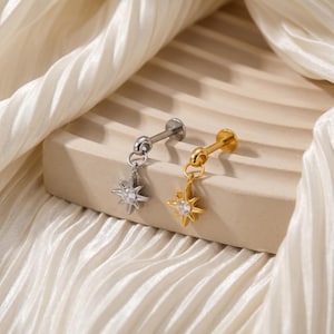 May include: Two star-shaped earrings, one silver and one gold, each with a small, clear gemstone in the centre. The earrings are displayed on a neutral-toned surface against a backdrop of soft, white fabric. The star pendants dangle from a small, circular attachment.