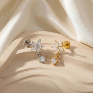 May include: Two flower-shaped earrings, one silver-toned and one gold-toned, each with a dangling round, clear gemstone. The flower petals are made of smaller, clear gemstones. The earrings are displayed on a beige surface.