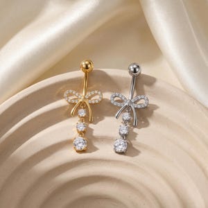May include: Two belly button rings, one gold-toned and one silver-toned, each featuring a bow design adorned with clear, sparkling crystals. The rings have a dangling three-stone design, with the stones getting progressively larger.