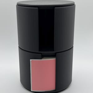 Puede incluir: Un dispensador de plástico negro con una pegatina rosa en el interior. El dispensador tiene una parte superior redonda y una forma cilíndrica.