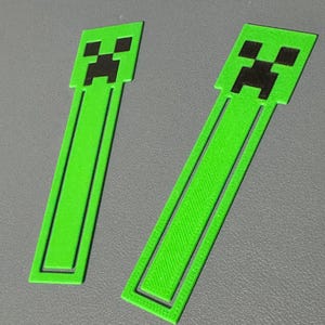 May include: Two green bookmarks with a pixelated black face design. The bookmarks are rectangular with a green border and a solid green centre. The design is inspired by a popular video game character.