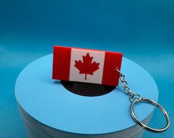 May include: A rectangular keychain featuring the Canadian flag. The flag design includes a red maple leaf on a white square, set against a red background. The keychain has a silver-colored chain and ring.