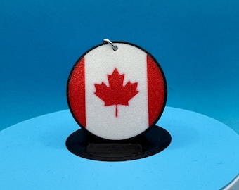 May include: A round charm featuring the Canadian flag, with a red maple leaf on a white background, framed by red borders. The charm has a silver loop for attachment and sits on a black stand against a blue backdrop.