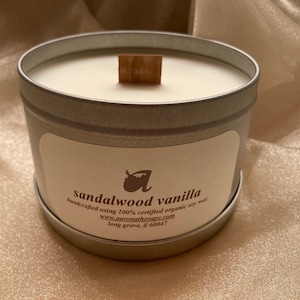 May include: A round, silver-colored candle tin with a wooden wick. The candle is filled with a creamy white wax. The label reads "sandalwood vanilla" and "handcrafted using 100% certified organic soy wax".