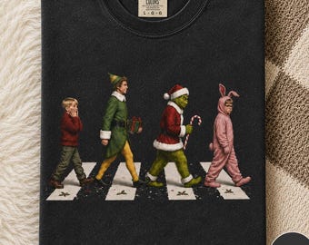 Op de afbeelding: Een zwart t-shirt met ronde hals en een afbeelding van vier figuren die over een zebrapad lopen. De figuren zijn een jongen in een rode trui, een elf in een groene jas, een groen wezen in een kerstmanpak en een kind in een roze konijnenpak.