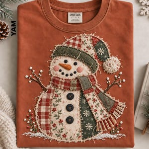 May include: A rust-colored crewneck t-shirt featuring a large, central applique of a snowman. The snowman is constructed from various fabric patches including red plaid, green herringbone, and floral prints, with three black buttons and a pom-pom hat.