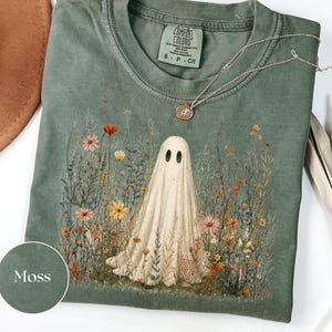 May include: A moss green crew neck t-shirt featuring a graphic of a white ghost standing in a field of colorful wildflowers. The shirt has a Comfort Colors brand label at the inner neckline and is paired with a delicate gold-toned chain necklace.