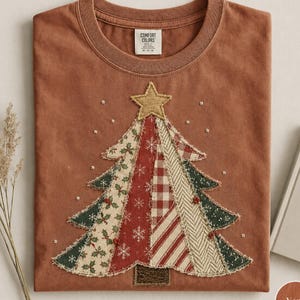 May include: A rust-colored t-shirt featuring a central Christmas tree graphic made of various fabric patches in red, green, and white patterns. The tree is topped with a gold star and accented with small white embroidered dots resembling snow.
