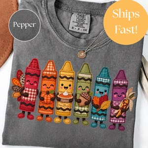 May include: A charcoal gray t-shirt featuring a graphic of six anthropomorphic crayons in red, orange, yellow, green, blue, and purple. Each crayon character holds a fall-themed item including a turkey, pumpkin, pie, acorn, leaves, and corn.