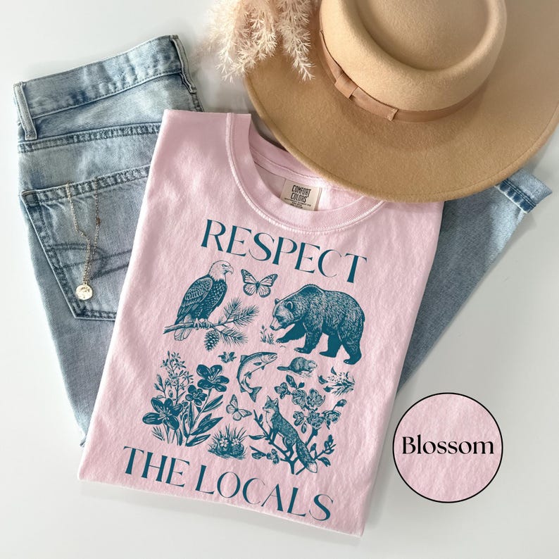 Outdoorsy nature graphic tee featuring North American wildlife artwork and “Respect The Locals” lettering, shown in a styled flat lay with denim jeans and a tan hat on a Blossom Comfort Colors Shirt.