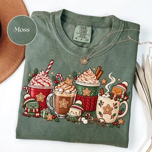 Folded Moss green Comfort Colors shirt featuring Christmas hot cocoa mugs, whipped cream, gingerbread cookies, candy canes and cheerful book characters reading in winter hats and scarves. Styled with a silver necklace and warm neutral accessories.