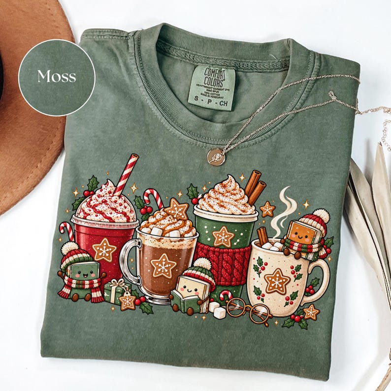 Folded Moss green Comfort Colors shirt featuring Christmas hot cocoa mugs, whipped cream, gingerbread cookies, candy canes and cheerful book characters reading in winter hats and scarves. Styled with a silver necklace and warm neutral accessories.