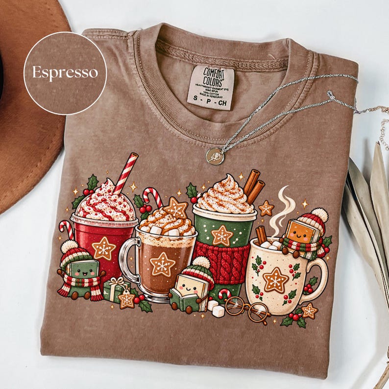 Folded Espresso brown Comfort Colors shirt displaying festive hot chocolate mugs surrounded by gingerbread cookies, candy canes, marshmallows, holly and adorable book characters wearing knitted winter hats and scarves. Styled with a silver necklace.
