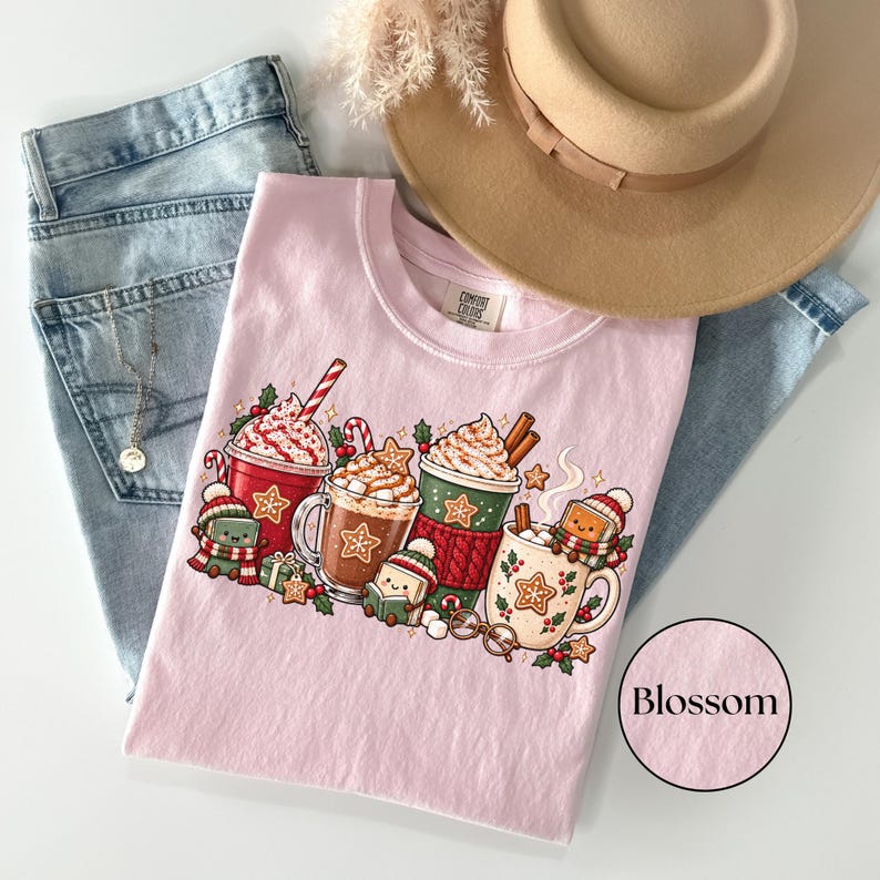 Folded Blossom pink Comfort Colors shirt featuring cozy Christmas hot chocolate mugs, whipped cream, candy canes, gingerbread cookies and adorable book characters reading in winter hats and scarves. Styled with jeans, pampas grass and a tan hat.
