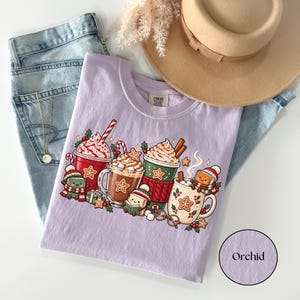 Folded Orchid lavender Comfort Colors shirt with a festive winter reading design featuring hot cocoa mugs, whipped cream, candy canes, gingerbread cookies and smiling book characters dressed in cozy scarves and knitted hats.