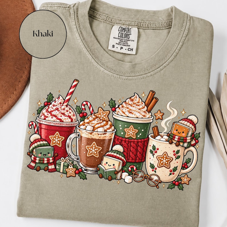 Folded Khaki Comfort Colors shirt featuring decorated Christmas hot cocoa mugs, whipped cream, gingerbread cookies, candy canes, holly and cute book characters reading in winter hats and scarves. Styled with a brown hat and neutral accessories.
