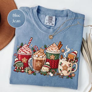 Blue Jean: Folded Blue Jean Comfort Colors shirt featuring a colorful Christmas reading design with festive hot cocoa mugs, whipped cream, candy canes, gingerbread cookies and cute book characters wearing winter hats and scarves. Styled with a gold necklace and warm neutral accessories.