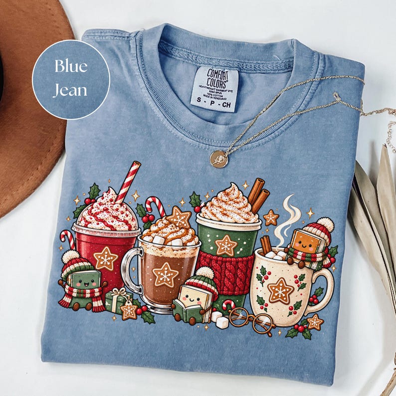 Blue Jean: Folded Blue Jean Comfort Colors shirt featuring a colorful Christmas reading design with festive hot cocoa mugs, whipped cream, candy canes, gingerbread cookies and cute book characters wearing winter hats and scarves. Styled with a gold necklace and warm neutral accessories.