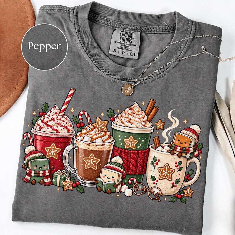 Folded Pepper gray Comfort Colors shirt featuring festive hot cocoa mugs, whipped cream, candy canes, gingerbread cookies and smiling book characters dressed in cozy scarves and winter hats. Styled with a delicate necklace and neutral holiday accessories.