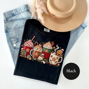 Folded Black Comfort Colors shirt featuring bright Christmas hot cocoa mugs, whipped cream, candy canes, gingerbread cookies, holly and cute book characters reading in striped hats and scarves. Styled with light-wash jeans, a gold necklace and a tan felt hat.