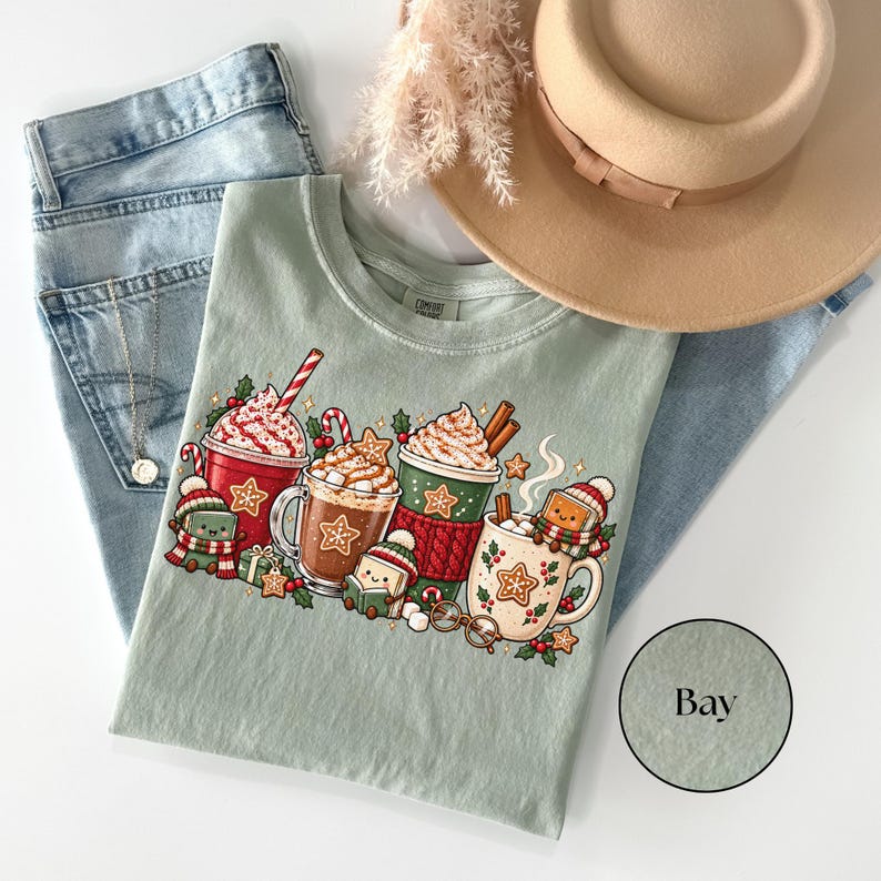 Folded Bay green Comfort Colors shirt with a cozy Christmas reading design featuring hot chocolate mugs, whipped cream, gingerbread cookies, candy canes and smiling book characters wearing winter hats and scarves. Styled with jeans and a tan hat.
