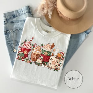 Folded White Comfort Colors shirt featuring festive red, green and cream hot cocoa mugs with whipped cream, gingerbread cookies, candy canes and small book characters dressed in knitted winter hats and scarves. Styled with jeans and a tan felt hat.