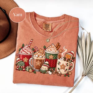 Folded Yam orange Comfort Colors shirt with a cozy Christmas book-lover design featuring decorated hot cocoa mugs, whipped cream, marshmallows, candy canes, gingerbread cookies and tiny book characters reading in winter hats and scarves.