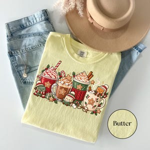 Folded Butter yellow Comfort Colors shirt with a festive book-lover illustration of hot cocoa mugs, whipped cream, candy canes, gingerbread cookies and small book characters dressed for winter. Styled with light-wash jeans, pampas grass and a tan felt hat.