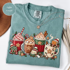 Folded Light Green Comfort Colors shirt with festive hot chocolate mugs, whipped cream, marshmallows, candy canes, gingerbread cookies and little book characters dressed for winter. Styled with a delicate necklace and neutral accessories.