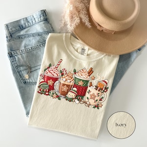 44.png — Denim: Folded Denim navy Comfort Colors shirt featuring a bright Christmas reading illustration with red, green and cream hot cocoa mugs, whipped cream, candy canes, gingerbread cookies and adorable book characters wearing winter hats and scarves.