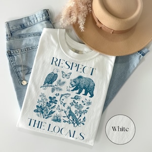 Nature lover graphic shirt featuring woodland wildlife and wildflowers in a detailed blue illustration beneath the phrase “Respect The Locals” on a White Comfort Colors T-shirt.