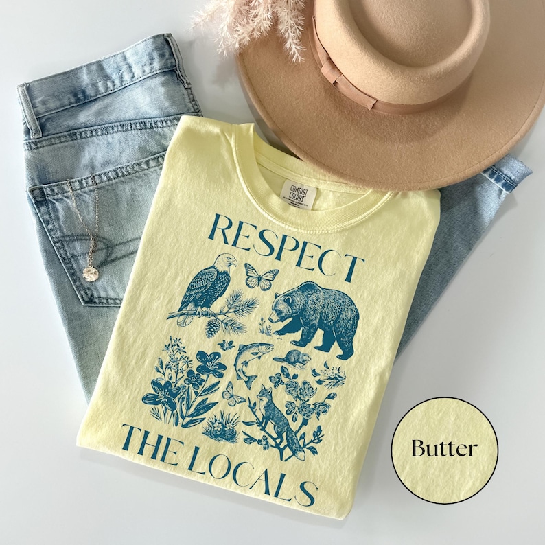Butter yellow Comfort Colors graphic T-shirt featuring a blue vintage wildlife illustration with a bear, eagle, fox, fish, butterflies, wildflowers, and the phrase “Respect The Locals.” Styled flat lay with light wash jeans, tan hat, and pendant necklace.