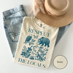 Ivory Comfort Colors wildlife graphic tee displaying a bear, eagle, fox, trout, butterflies, pine branch, and floral artwork with “Respect The Locals” typography.