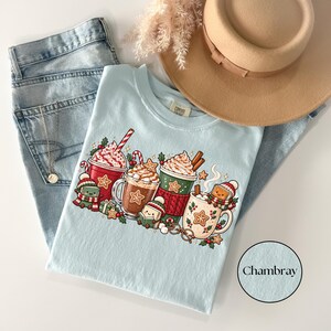 Folded Chambray blue Comfort Colors shirt displaying festive hot cocoa mugs, whipped cream, candy canes, gingerbread cookies, holly and cheerful book characters wearing winter scarves and hats. Styled with light-wash jeans and a tan felt hat.
