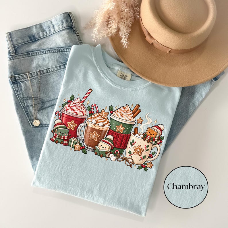 Folded Chambray blue Comfort Colors shirt displaying festive hot cocoa mugs, whipped cream, candy canes, gingerbread cookies, holly and cheerful book characters wearing winter scarves and hats. Styled with light-wash jeans and a tan felt hat.