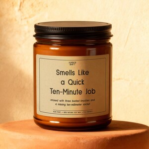 May include: A brown glass jar candle with a black lid sits on a terracotta-colored stand. The candle has a label that reads "Smells Like a Quick Ten-Minute Job." The background is a warm, neutral tone with a sprig of greenery.